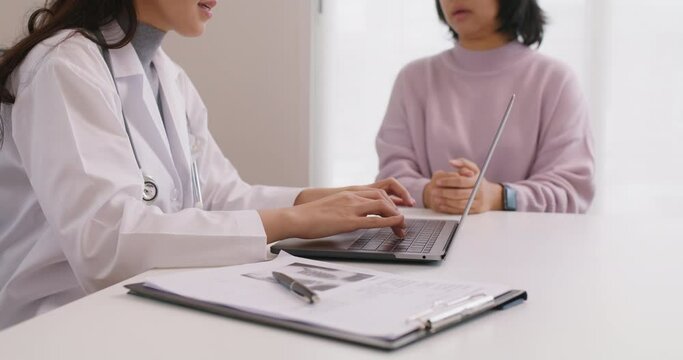 Adult female asia illness people see visit GP at clinic office. Medic health care sit work on desk talk in exam result, ask sick symptom, give advice help, typing Rx pill enter data on laptop app.