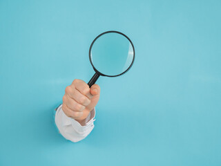 A woman's hand sticking out of a hole in a paper blue background holds a magnifying loupe. 