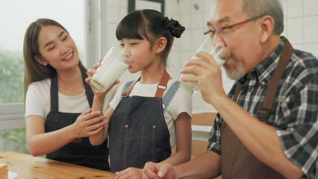 Asian  family enjoying breakfast at cozy kitchen, little girl daughter sitting on table, drinking milk with smiling father and mother in morning. Happy family in kitchen.