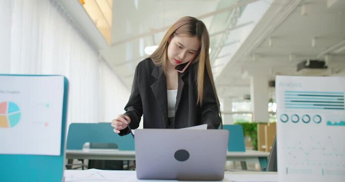 Young busy asian business woman working on project paperwork talking phone, using laptop remotely work in modern office. Female freelancer entrepreneur making distance job telesale call with customer.