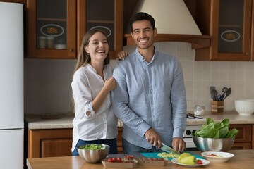 Cheerful wife and husband smile look at camera enjoy cooking vegetarian salad or vegan dish for romantic date pose in cozy kitchen at home. Married couple prepare natural healthy food together concept