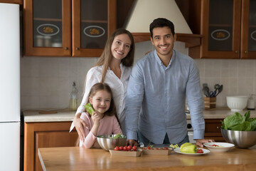 Fototapeta premium Parents with little adorable daughter posing standing in cozy domestic kitchen at home smiling look at camera feel happy, cooking together vegetarian dish. Family recipe, healthy natural eat concept