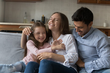Young attractive parents and preschool adorable daughter play seated on couch, laughing, feeling overjoyed, tickling each other enjoy playtime and carefree active family leisure having fun at home