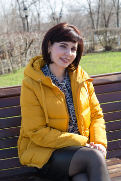 An adult brunette woman in a yellow jacket smiles gently sitting on a bench in the park.