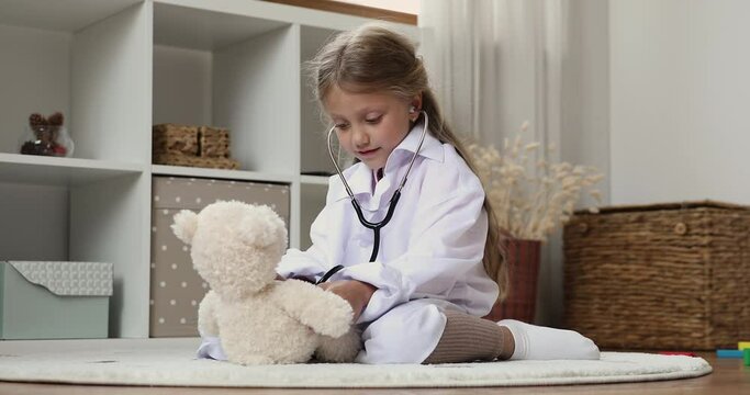 Engaged cute preschool girl in oversized white coat examining toy with stethoscope, listening to teddy bear patient heartbeat, sitting on heating floor in playroom, enjoying game