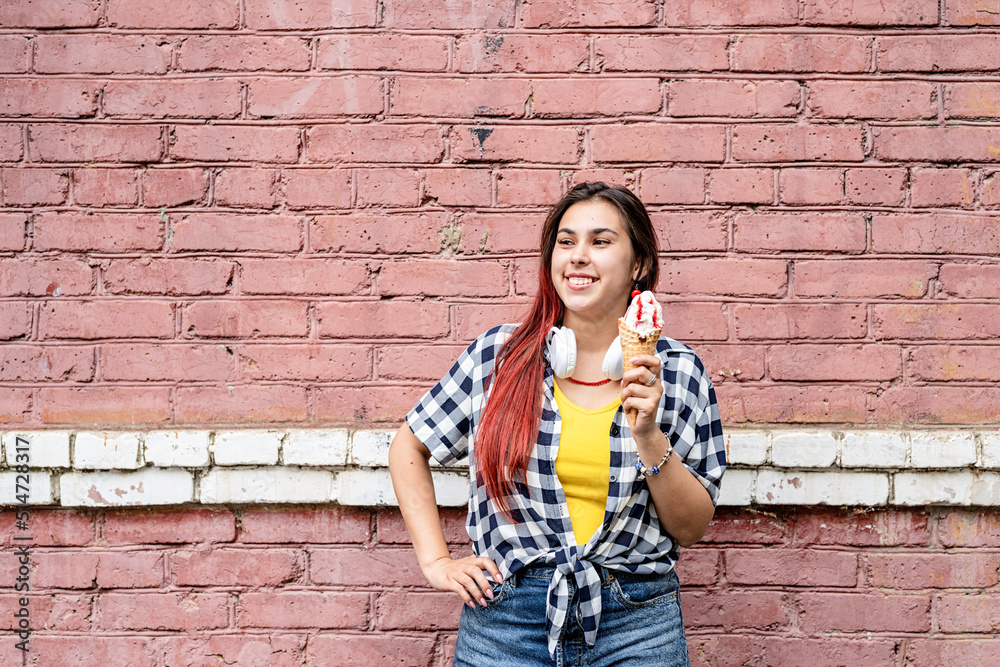 Wall mural cheerful trendy woman with red hair eating ice cream on pink brick wall background at street