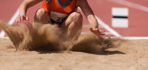 Female athlete long jump landing sand spray, landing in long jump in track and field - obrazy, fototapety, plakaty
