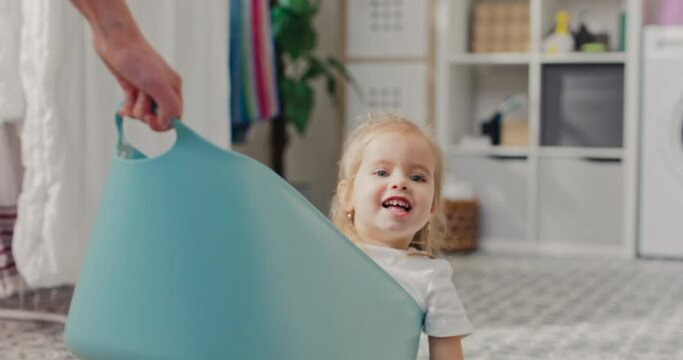 Close-up of face of cute little blonde girl sitting in clothes bowl. Woman pulls girl for fun mom spends time with daughter in laundry room.