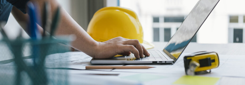Young man with a laptop plotting a system of building structures in blueprints, Architects or engineers are designing buildings using computers to calculate the physical structure to be correct.