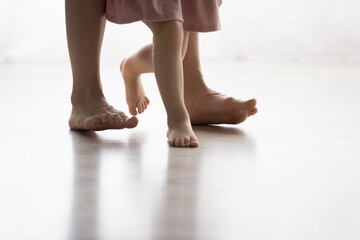 Barefoot feet of unrecognizable caring mother and little preschool daughter in dress walking on warm floor with underfloor heat system at modern home. Podiatry, childcare, support, motherhood concept