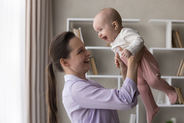 Obraz premium Young 25s beautiful mother lifts holds in arms playing with adorable newborn daughter, cute baby girl, family having fun enjoy playtime at home. Happy motherhood, carefree babyhood, maternity concept