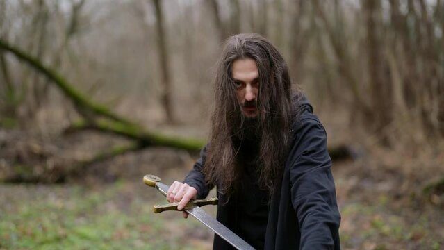 portrait of a bearded man with long hair and dark clothes against a background of blurred moss-covered trees. he looks into the camera, holds the sword and keeps the blade with hand