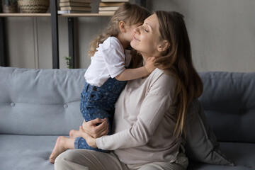 Preschool 4s lovely daughter hugs and kisses her young attractive pregnant mother, child express caress feeling love sit together on sofa at home. Happy maternity, motherhood, family bonding concept