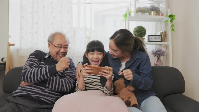 Asian happy family play time. little girl with grandparents together playing video games use joystick control enjoy laughing fun in living room home at night. Happy child happiness lifestyle concept.