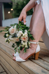 Obraz na płótnie Canvas the wedding bouquet is in the bride's hand and lowered to her feet. The bride's legs and arms, her shoes and part of her dress are visible. She is sitting on a wooden bench