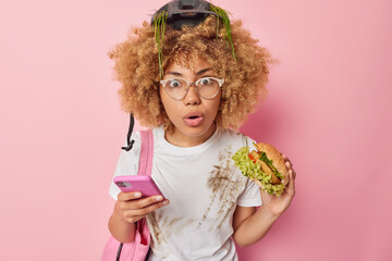 Shocked teenage girl with curly hair has delicious lunch eats hamburger holds mobile phone sends text messages cannot believe own eyes wears spectacles casual white t shirt isolated over pink wall