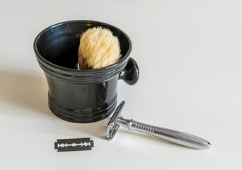 a boar bristle shaving brush in a foam cup next to a t-shaped shaving machine and a replacement razor on a white table.