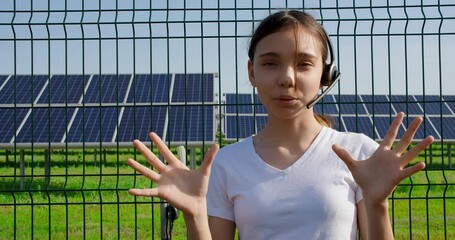 A teenage girl with a headset is standing near a solar panel farm and is actively gesticulating on her online streaming about sustainable energy production.