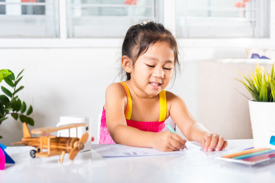 Asian cute kid preschooler sitting on table smiling she draw and writing picture with pencil at home to learning arts homework, Happy child little girl drawing cartoon on paper before paint the color
