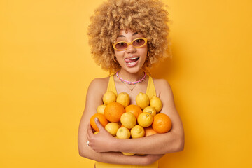 Positive curly haired young woman wears sunglasses and t shirt embraces heap of citrus fruits...