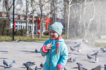 Laughing girl in a spring park. A child with long hair braided into pigtails. A girl in a blue jacket. A flock of pigeons is in the background. Childhood.  Daytime.