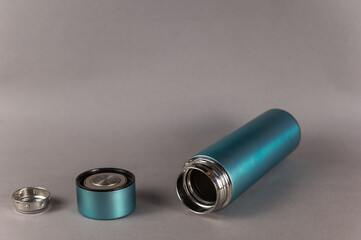 Modern thermos, lid and teapot against a gray background. Cylindrical vacuole flask in turquoise color. Container for hot and cold drinks. Equipment for sports, hiking, travel.