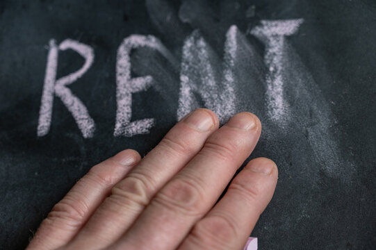 The semi-erased word RENT on the black chalkboard. An adult man's left hand removes the handwritten word with his fingers. Fuzzy letters on black surface. - Powered by Adobe