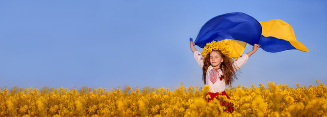 Ukrainian child girl in embroidered shirt and yellow wreath in field of yellow flowers against blue sky. Ukrainian blue-yellow flag flying in wind in hands of little Ukrainian girl. Banner.