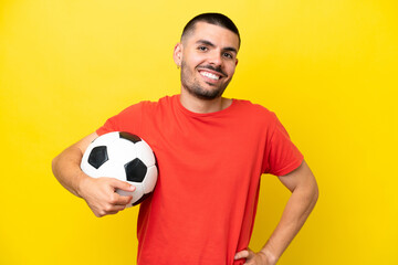 Young caucasian man playing soccer isolated on yellow background posing with arms at hip and smiling