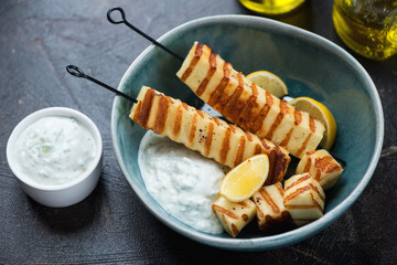 Green bowl with halloumi kebabs and tzatziki dip sauce on a dark-brown stone background, studio shot