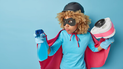 Indoor shot of curly woman cleaner pretends being super hero holds spraying bottle of detergent and...