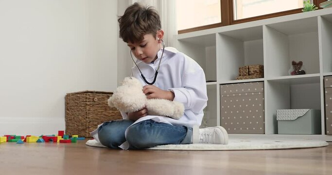 Focused sweet preschool boy wearing medical white coat, playing doctor at home, examining teddy bear with stethoscope, holding, listening to toy patient. Childhood, medicine, future profession concept