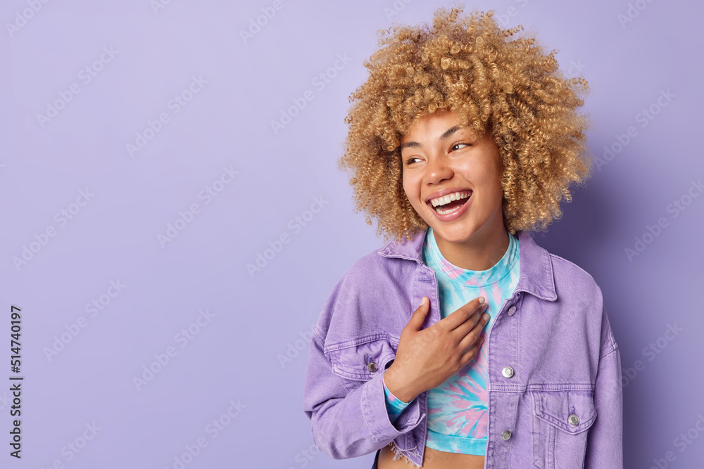 Wall mural Photo of positive curly haired European woman keeps hand on chest laughs gladfully dressed in top and jacket cannot stop laughing isolated over purple background copy space for your promotion