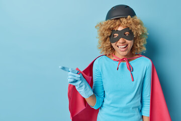 Cheerful female superhero smiles happily points away on blank space prepares for masquerade stands...