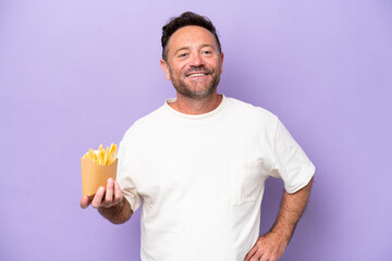 Middle age caucasian man holding fried chips isolated on purple bakcground posing with arms at hip and smiling