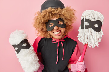Cheerful curly haired woman poses with cleaning tools holds mops going to wash floor wears helmet...