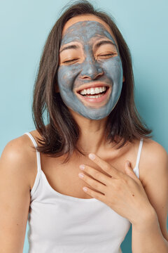 Overjoyed Asian Woman Has Dry Skin Applies Nourishing Clay Mask For Moisturising Smiles Broadly Keeps Hand On Chest Tries To Improve Symptoms Of Dermatitis Wears White T Shirt Isolated On Blue Wall