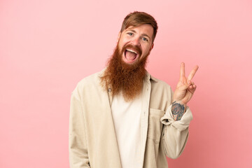 Young reddish caucasian man isolated on pink background smiling and showing victory sign