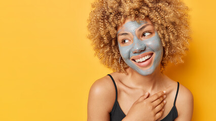 Horizontal shot of positive curly haired woman applies nutrient clay mask on face to detoxify skin smiles gladfully dressed in casual t shirt isolated over yellow background blank space for promo