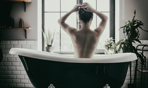 Graceful Woman Is Sitting On The Bathtub In The Bathroom In Front Of The Window