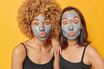 Indoor shot of surprised young women apply beauty clay mask on face undergo skin care procedures dressed in casual black t shirts look wondered at camera isolated over vivid yellow background