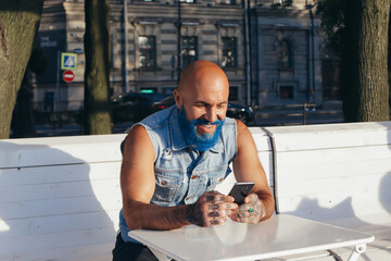 fashionable man with a blue beard sits at a white table and uses a mobile phone © Елена Вырыпаева