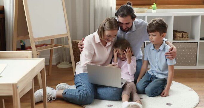 Happy young parents and two little kids using watching movie on laptop, smiling, laughing. Family couple hugging children, relaxing on heating floor in playroom, using computer. Family leisure concept
