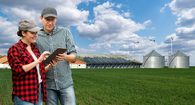 Two farmers with tablet computer on a background of modern dairy farm using renewable energy, solar panels and wind turbines
