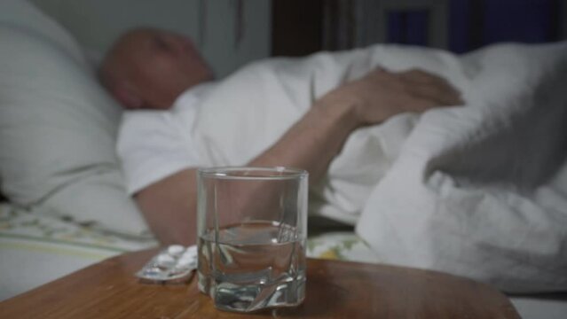 Nightstand with pills and glass of water stands near bed with older sick man turning in bed of home or nursing house