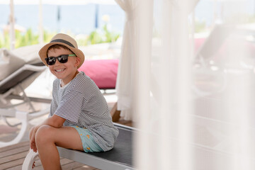 Smiling Caucasian boy in straw hat and sunglasses having rest sitting on chaise lounge on the beach. Vacation in luxury resort hotel. Copy space.