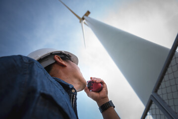 Asian electrical engineer man using  radio communication, with blur of Wind turbines background, to...