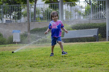 Little boy playing with garden sprinkler. Preschooler Boy running and jumping. Summer outdoor water fun in the backyard. Children play with hose watering grass. Kids run and splash on hot sunny day.