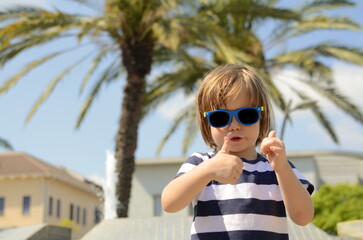 Funny kid in sunglasses on the background of palm trees. Concept: family resort, traveling with children, exotic vacation. Bright sunny day, good mood, positive little boy in a striped t-shirt