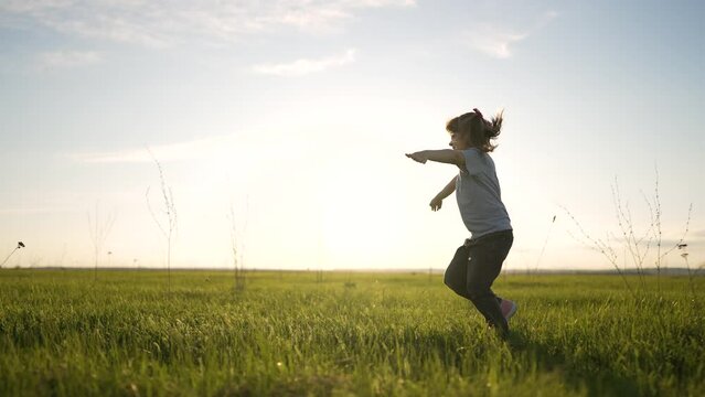 Happy child run her arms to sides. Girl play in park at sunset. Little girl run through green grass in the field. Dream girl. Cheerful child runs smiling and dreaming. Winner girl strives for success